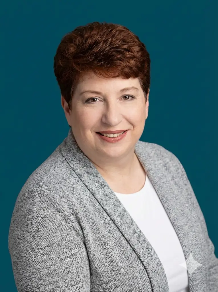 Photo shows Laura, a white woman with dark red hair and brown eyes. She has on a gray sweater over a white blouse against a dark blue background.