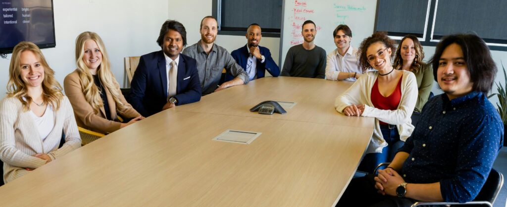 A group of 10 people sitting around a rectangular table that has a phone conference device sitting in the middle of it. A couple of people were wearing suits, but most people are in business casual clothing. They are listening attentively to a person sitting at the head of the table.