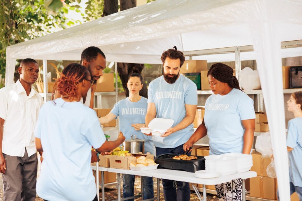 A photo showing a group of four people in light colored shirts serving food to an individual under a white canopy during a food distribution event.