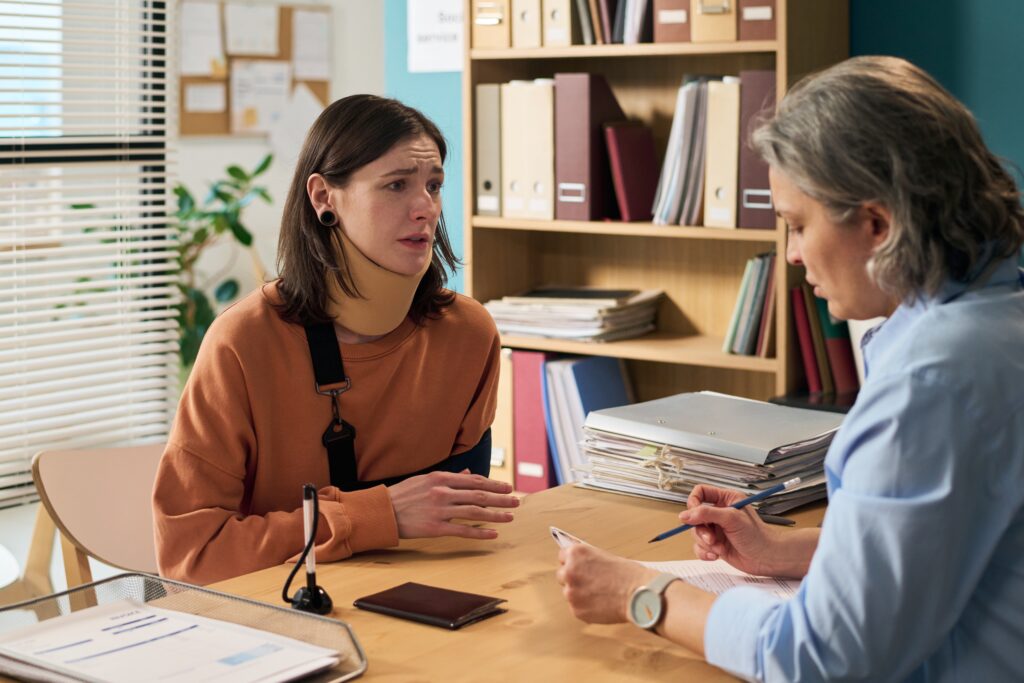 A photo featuring a female social worker meeting with a client in an office setting. They are sitting at a round table. The social worker has a pen and paper and there are files on the table. The client has a concerned look on her face.
