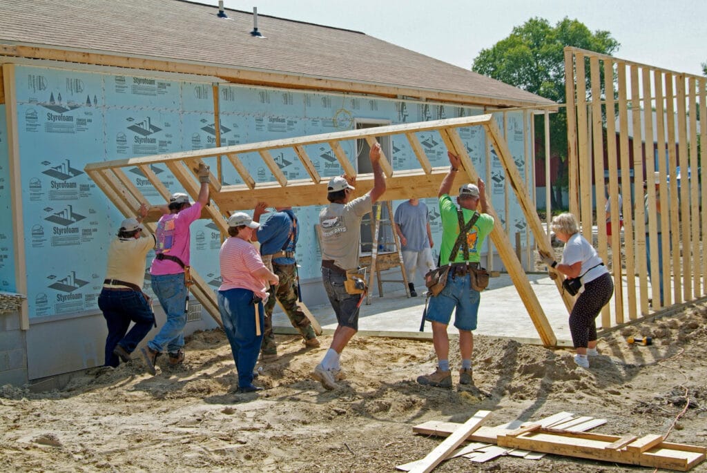A photo showing a group of eight men and women working at a construction site for a new home. They are all lifting a wooden frame into place. Some members of the group are dressed in casual clothing whiile others are wearing safety vests. There is a home in the background covered in blue tarp.