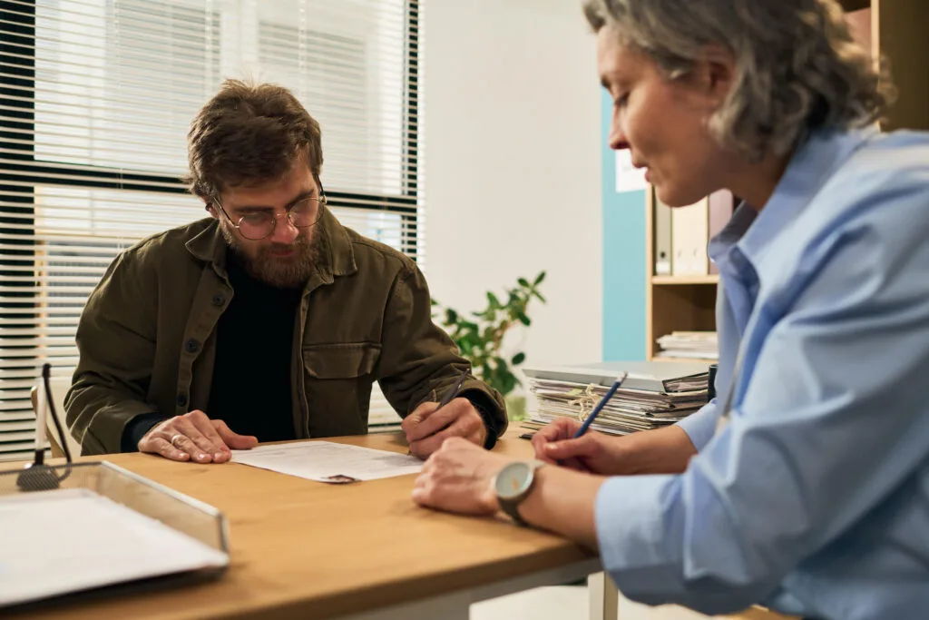 Home 5 A photo of a caseworker meeting with a man at an office desk. The caseworker watches as the man reviews and signs paperwork.